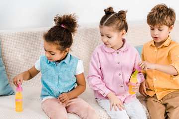 adorable multiethnic children sitting on sofa and blowing soap bubbles