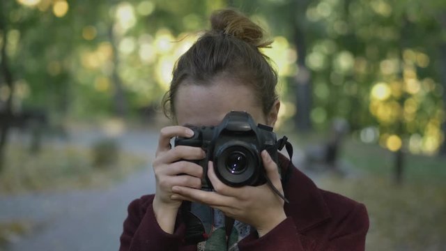 Professional young woman photographer with digital camera taking pictures at autumn city park. Close up, slow motion, shallow depth of field.