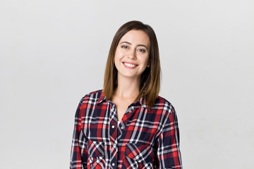Smiling positive female with attractive look, wearing checkered shirt, posing against white blank wall