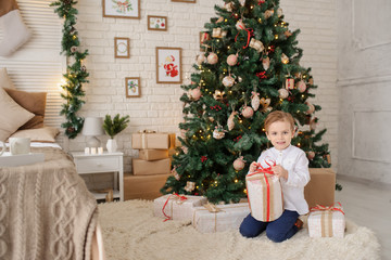 Obraz na płótnie Canvas a 6-year blond boy with a present in his hand near a christmas tree and a bed