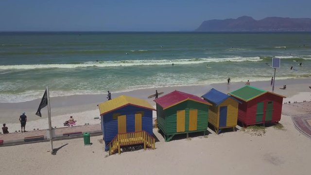 Colourful wooden beach huts with solar panel, shark warning flag and unidentifiable people surfing as seen from above on the beach at Muizenberg near Cape Town, South Africa.
