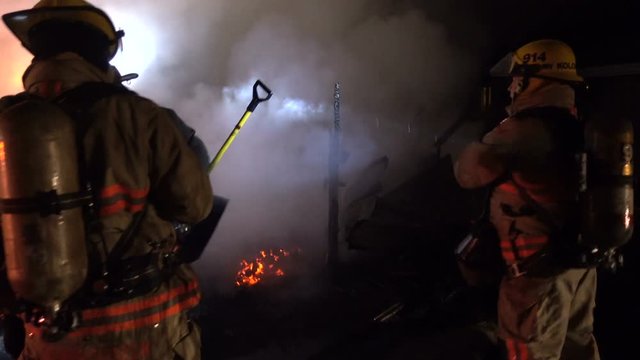 Firemen examine and spray smoking remains of burnt building. Fire is extinguished, but water is sprayed on the ashes by fire response team to snuff out embers that might relight. Rear view of firemen.