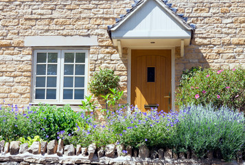 Light brown doors in a limestone golden colored English cottage with flowers and shrubs in a front...
