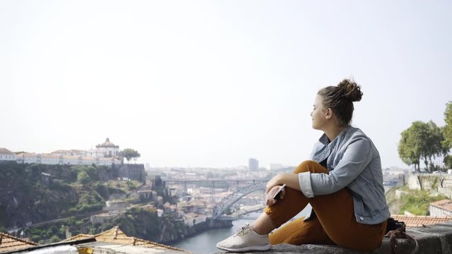 Young woman in casual wear enjoying beautiful cityscape sitting on high viewpoint above buildings with red tiled roofs and big river. Porto, Portugal