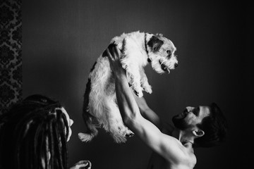 A man without a shirt is holding a dog on his outstretched arms. Nearby is a girl in dreadlocks.