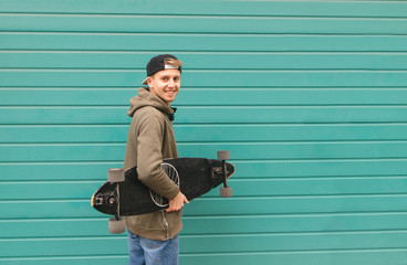 Happy student with a longboard in his hands on a color turquoise background, looking into the camera and smiling. Portrait of a positive skateboarder. © bodnarphoto