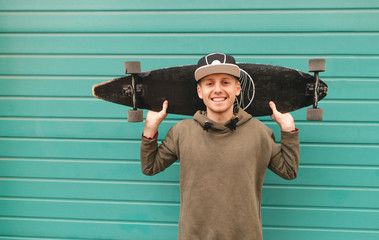 Smiling teenager in a cap and a hoodie stands on the background of a green wall, holds a longboard on his shoulder, looks into the camera and rejoices. Portrait of lucky skater. Copyspace