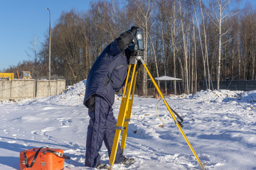 Surveyor assembles a geodetic instrument from an orange case and a yellow tripod at a construction site in winter
