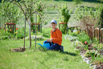 Cute European boy is helping his parents in the countryside croft. He is sitting under the tree in the garden with watering can in his hands and looking toward the camera.