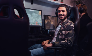 A young cheerful Indian guy wearing a military shirt sitting on a gamer chair and looking at a camera in a gaming club or internet cafe.
