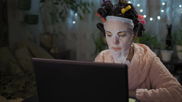 Beautiful Woman in Rejuvenating Mask and Hair Curlers Looks at Laptop Screen