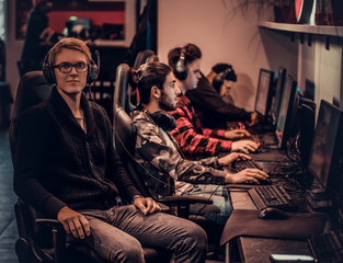 A young smart gamer wearing a sweater and glasses sitting on a gamer chair and looking at a camera in a gaming club or internet cafe.