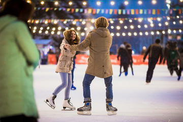 Young couple in love Caucasian man with blond hair with long hair and beard and beautiful woman have fun, active date skating on ice scene in town square in winter on Christmas Eve