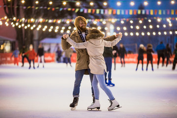 Naklejka premium Young couple in love Caucasian man with blond hair with long hair and beard and beautiful woman have fun, active date skating on ice scene in town square in winter on Christmas Eve
