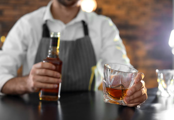 Bartender with glass and bottle of whiskey at counter in bar, closeup. Space for text