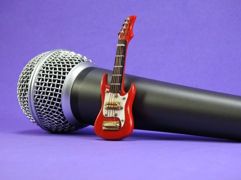 A red miniature electric guitar propped up on a dynamic handheld vocal microphone. Purple background.