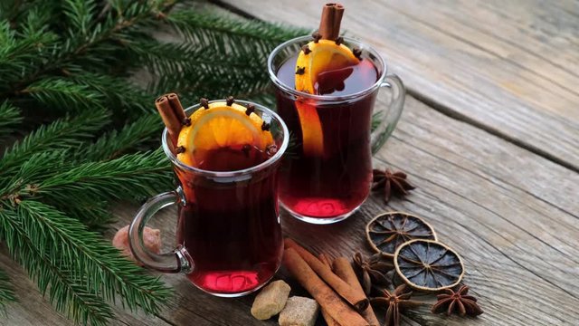Glass mugs of mulled wine with spices, citrus fruits and Christmas tree branches. Lights of a fireplace flame or lights of a Christmas fir tree are reflected on glass mugs.