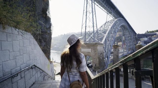 Young woman walking on street under bridge Ponte Luis I in old city centre Porto. Following girl in dress and hat walking down on stairs at background historic bridge