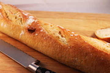 Close-up of a french baguette on a wooden cutting board. 1 slice has been cut from the baguette, the knife is lying beside it on the cutting board