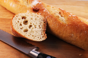 Close-up of a french baguette on a wooden cutting board. 1 slice has been cut from the baguette, the knife is lying beside it on the cutting board