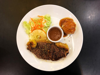 Top view of Serving size of Beef Steak and slice pineapple, brown sauce cup in white dish on black wooden table.