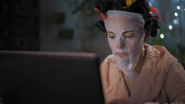 Close-Up, Beautiful Woman Working on Laptop in Rejuvenating Mask on Face and in Curlers