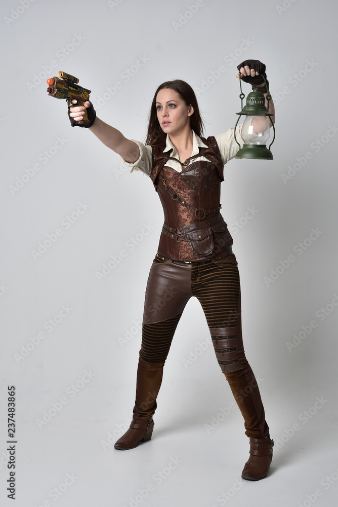 Poster  full length portrait of brunette  girl wearing brown leather steampunk outfit. standing pose holding a gun on grey studio background.