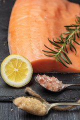 Raw salmon fillet on black slate plate and ingredients for making gravlax. Salt, sugar, lemon and rosemary in the background for dry cure marinade, dark background.