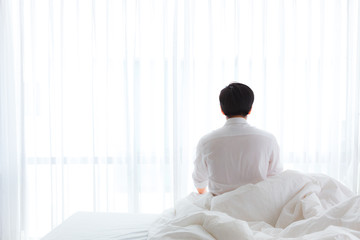 Black hair of young business asian man in casual white shirt sitting near the big white window after waking up with sunrise at morning, back view.