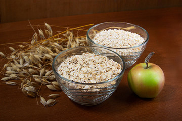 Oat ears stems and oat flakes and green apple in a bowl on a dark brown wood background. oat flakes big and small size grind. oatmeal flakes coarse. large-sized flakes. Useful fiber-rich product.