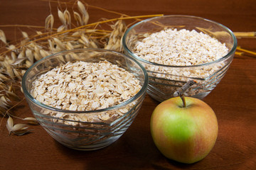 Oat ears stems and oat flakes and green apple in a bowl on a dark brown wood background. oat flakes big and small size grind. oatmeal flakes coarse. large-sized flakes. Useful fiber-rich product.