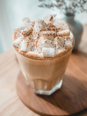One glass of hot coffee with marshmallow on top serve on white tray on wood table next to bright window in morning.