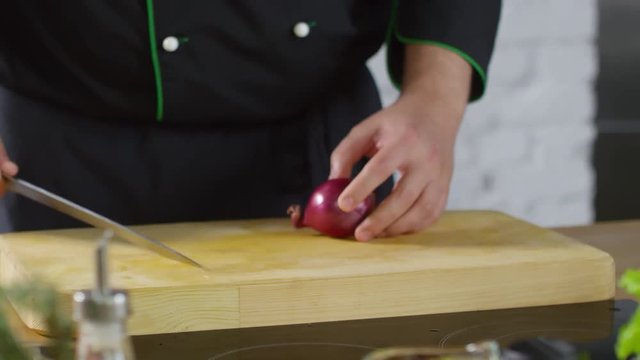 Mid-section tracking shot of unrecognizable male cook in uniform cutting red onion on wooden board
