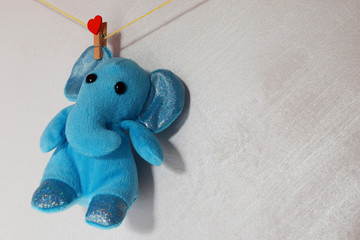 Blue cute plush toy elephant hanging on yellow rope with red heart shape peg on light grey wall background with copy space for text.