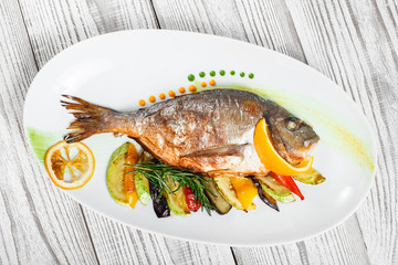 Grilled dorado fish with baked vegetables and rosemary on plate on wooden background close up. Healthy food. Top view