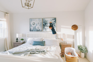 A little boy jumping in the air in a bright white bedroom.