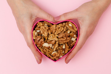 Woman's hand holding a box in heart shape with peeled walnuts on pink background.