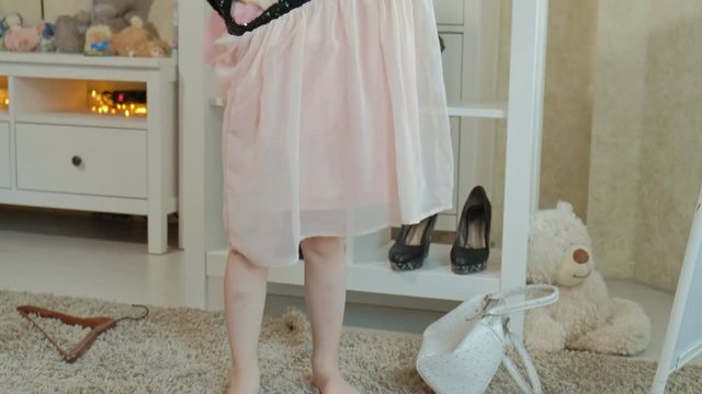 cheerful girl with pigtails in a pink skirt tries on adult mother's clothes, dresses, high heels and dances in front of a mirror with a bag, mothers wardrobe