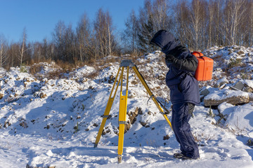 Surveyor carries a geodetic instrument in an orange case and a yellow tripod at a construction site in winter