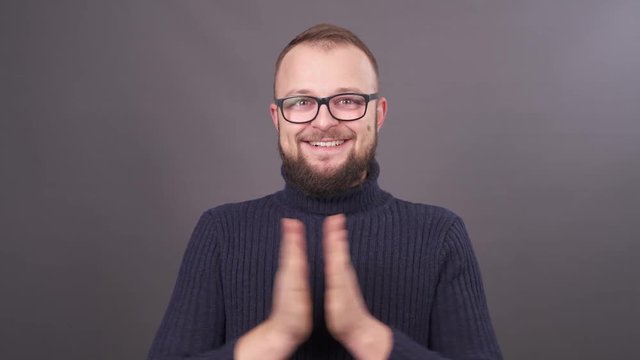 Close-up portrait of a bearded young caucasian man in glasses clapping his hands. Isolated on grey background.