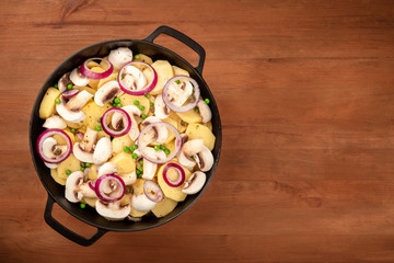A photo of a braiser with raw vegetables, potatoes, onions, green peas, and mushrooms, shot from the top on a dark rustic wooden background wih a place for text