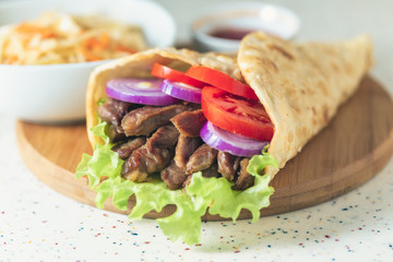 Handmade doner kebab is lying on the white table made of artificial stone Shawarma on the wooden cutting board with chicken meat, onions, salad lies on a dark old wooden table.