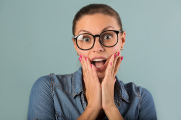 beautiful young girl in a denim shirt with glasses on a blue background with astonishment on her face