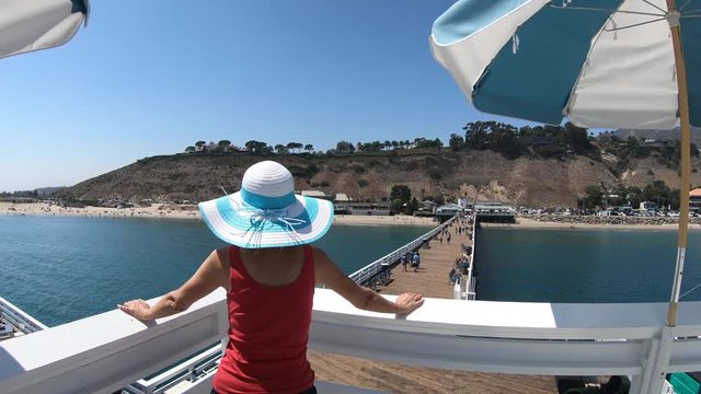Caucasian woman at Malibu Pier in California West Coast, United States. Blonde girl enjoys Santa Monica Mountains and Surfrider Beach. Aerial view over turquoise Pacific Ocean. Summer holidays.