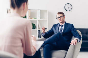 selective focus of handsome businessman in suit and glasses giving interview to journalist in office