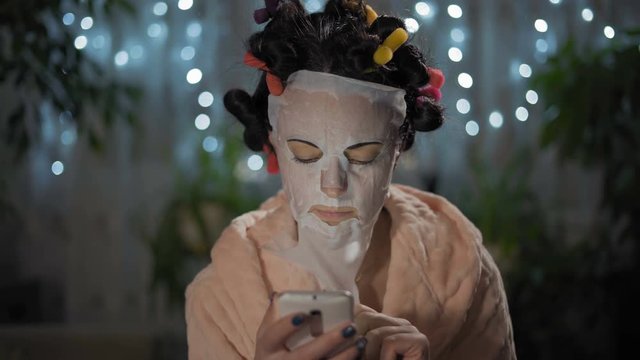 Close-up, Woman in Pink Robe Writes a Message on a Smartphone in a Rejuvenating Mask and Curlers
