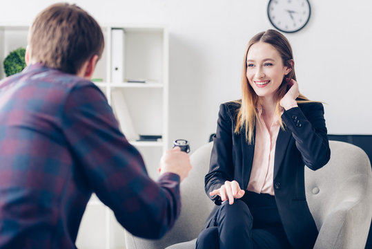 smiling businesswoman in suit touching hair while giving interview to journalist in office
