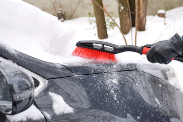 Grey car outside covered with snow and ice. man in blue jacket and leather black gloves shoveling snow from window glass and windscreen by red plastic brush. Winter season work.