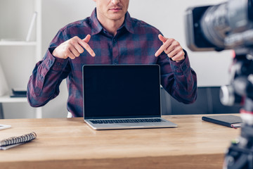 cropped image of male video blogger recording vlog and pointing on laptop with blank screen in office
