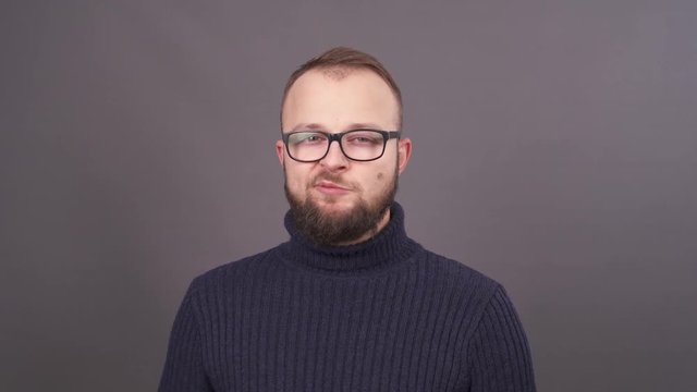 Close-up portrait of a bearded young caucasian man in glasses, shows at camera gums. Puts it into mouth and starts to chew and slurp. Isolated on grey background.